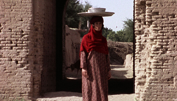 A woman is standing under the arch of a very old looking building. She is balancing a tray and bowl on her head.