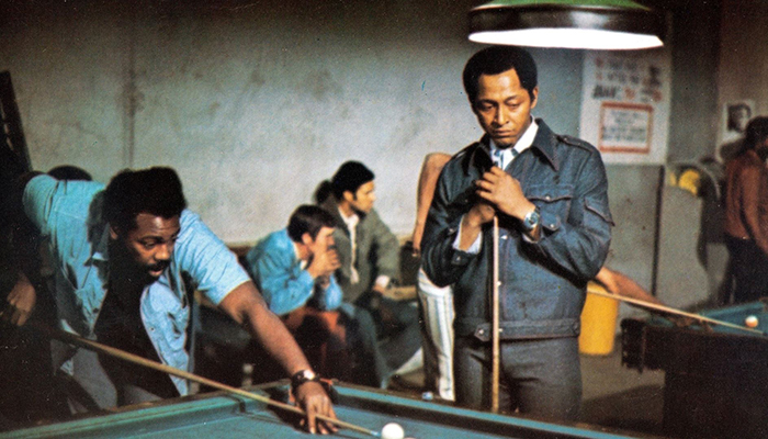 Two Black men playing pool in a pool hall. One man is bent over shooting, the other is standing nearby watching.