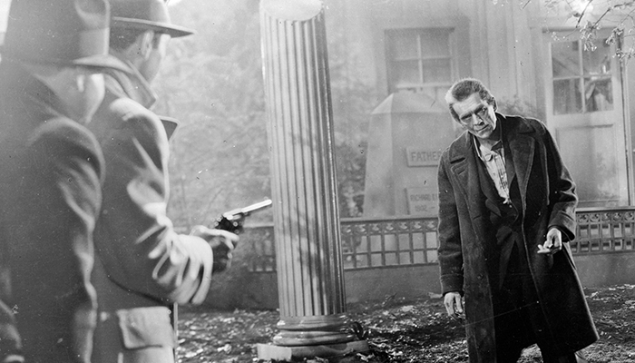 Two men stand in a cemetery pointing a gun at an approaching man.