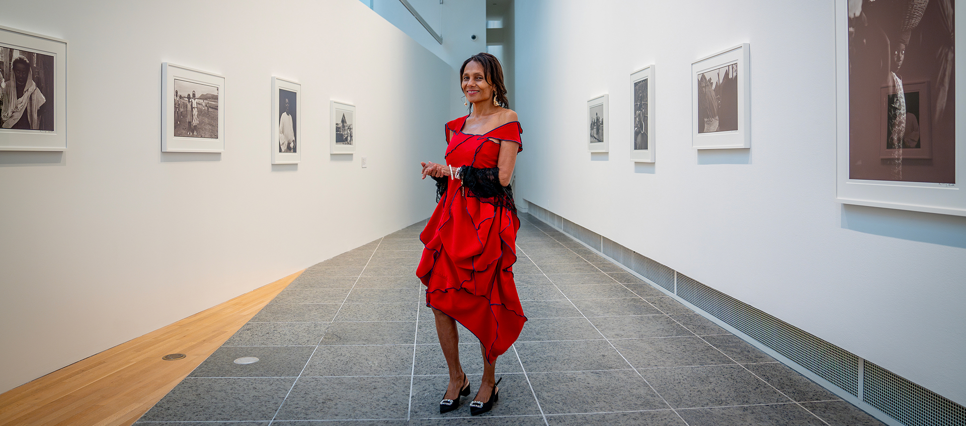 Ming Smith, wearing a red dress, stands in the middle of a narrow, triangular gallery space. Framed photographs hang on the walls on either side of her. 