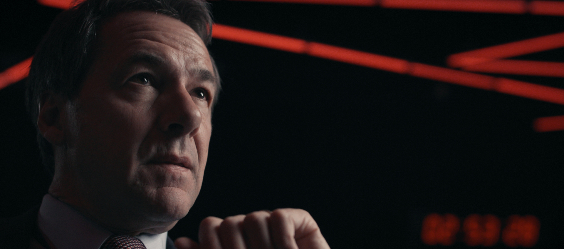 A closeup of a white, middle-aged man in a suit looking offscreen. He is in a dark situation room with dramatic red lines of lights on the ceiling.