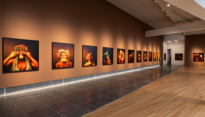 View of 14 large-scale color photographs on a brown gallery wall. The images show young Black men wearing or interacting with plants, fruit, or shells.