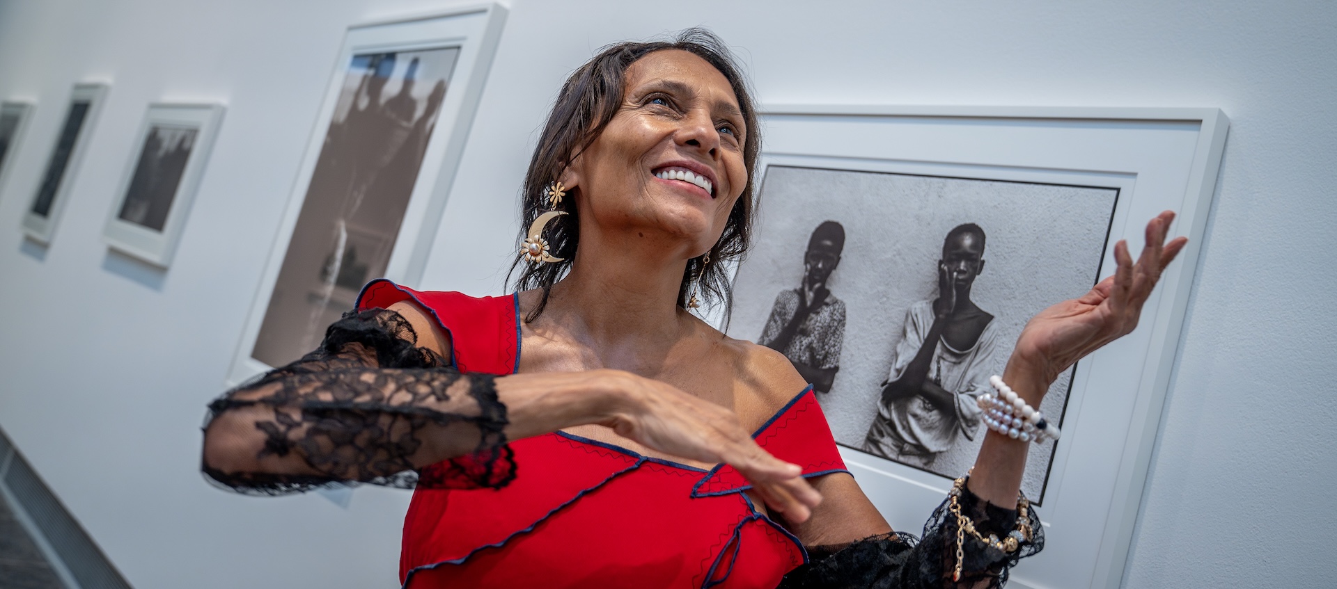 A woman in a red dress looks up and smiles in the middle of a gallery with walls holding framed photographs.