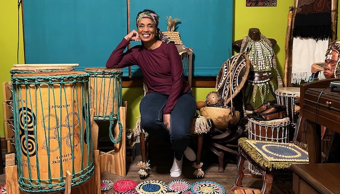 A smiling woman sits in her home surrounded by drums, chairs, and other handmade items.