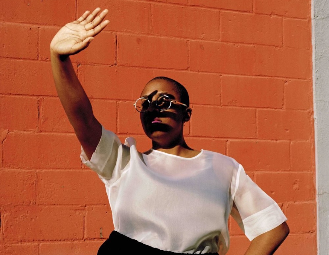 A woman stands with one hand on her hip and the other held out and up to shield her face from the sun.