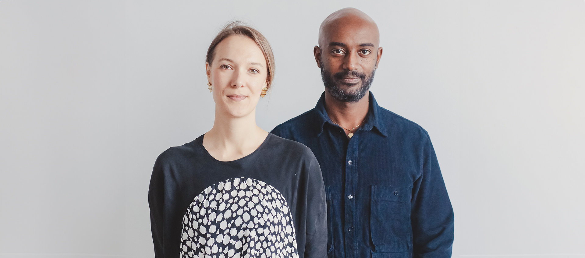 Jen Wood, who has pale skin and light-brown hair stands to the left of Emanuel Admassu who has medium brown skin, is bald, and has a short beard. 