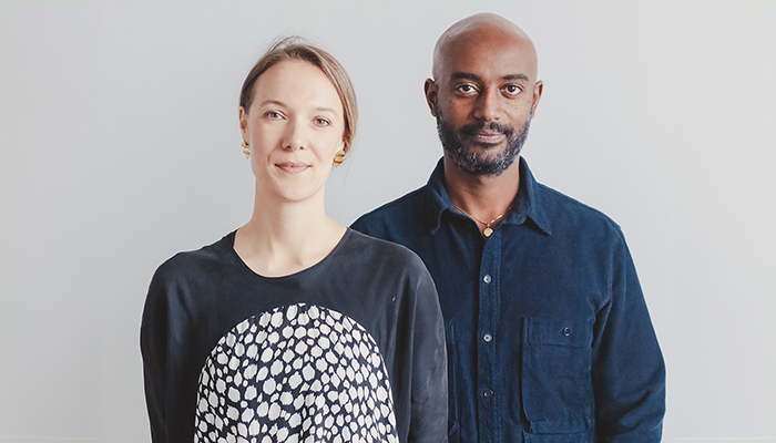 Jen Wood, who has pale skin and light-brown hair stands to the left of Emanuel Admassu who has medium brown skin, is bald, and has a short beard. 