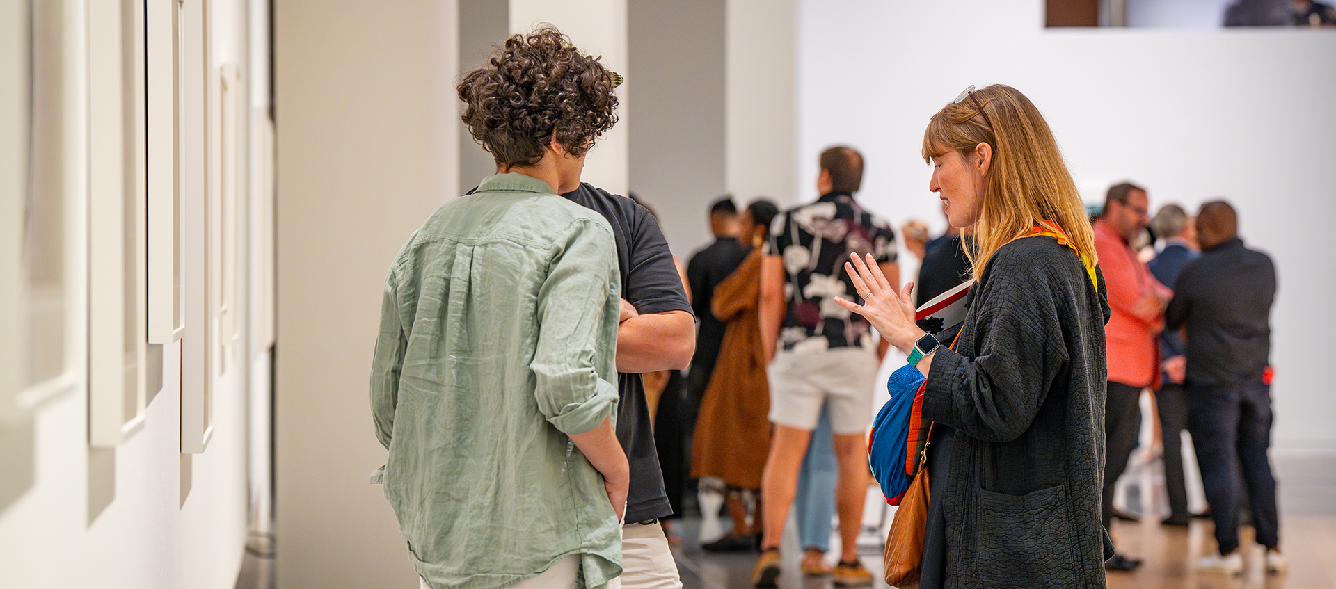 People stand in groups in a large gallery. In the foreground, three people have a conversation next to framed works hanging on the wall. 