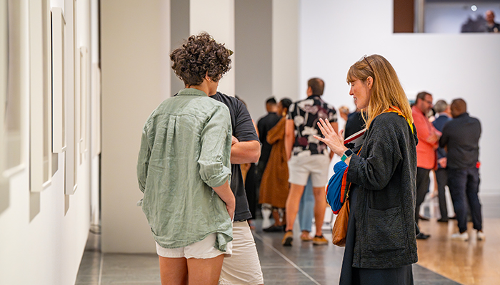 People stand in groups in a large gallery. In the foreground, three people have a conversation next to framed works hanging on the wall.