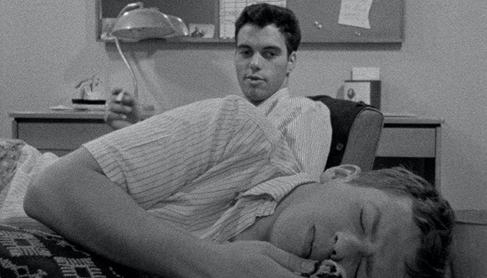 A young man is laying down and another young man is sitting behind him holding a cigarette. A desk, lamp, and bulletin board are in the background.