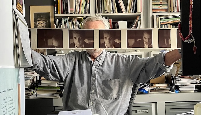 A man sits at a desk holding a horizontal printout of pictures of himself in front of his face.