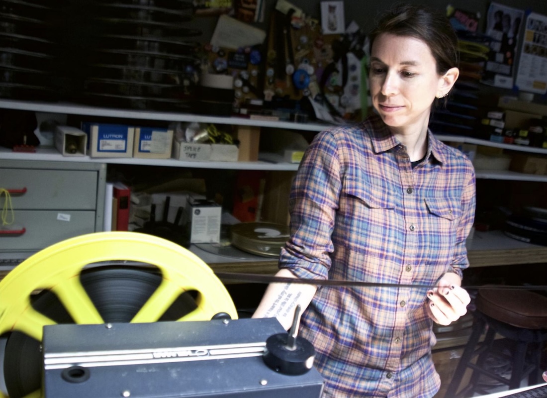 A woman stands in a projection booth, inspecting 35mm film unspooling off a reel.