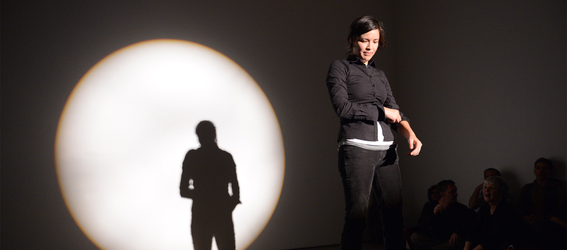 Maria Hupfield stands in a spotlight onstage wearing a black button-down shirt and jeans. Her silhouette is visible in a large circle of light behind her.