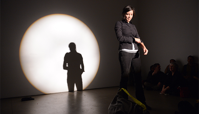 Maria Hupfield stands in a spotlight onstage wearing a black button-down shirt and jeans. Her silhouette is visible in a large circle of light behind her.