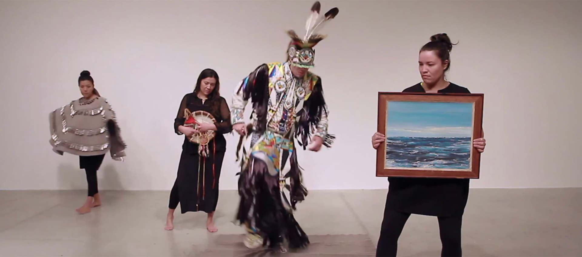 Four performers wear Indigenous ceremonial or black clothing. One wears a gray felt shawl with silver jingles. One holds a landscape painting.