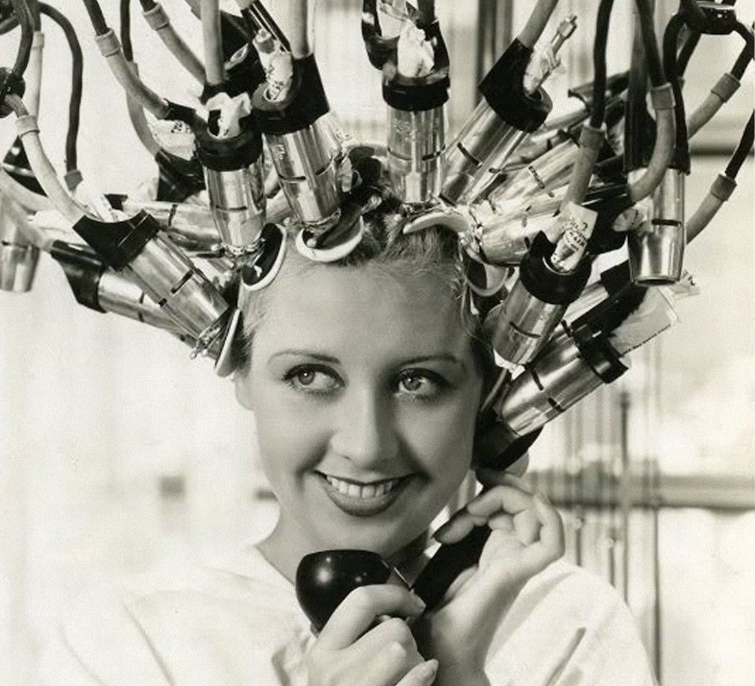 A woman with her hair in the tubes of a vintage salon styling tool smiles as she talks on the phone.