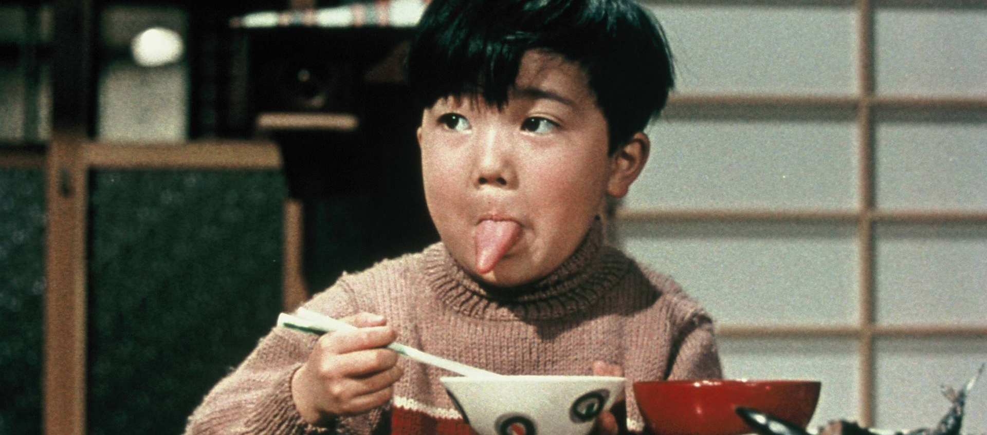 A young boy sits at a table holding chopsticks over a bowl with his tongue sticking out.