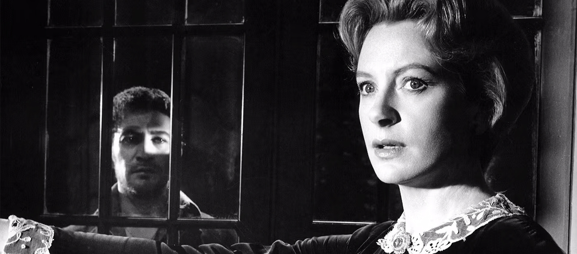 1.	A black and white closeup of a woman standing in front of a window with a man staring at her through the window.