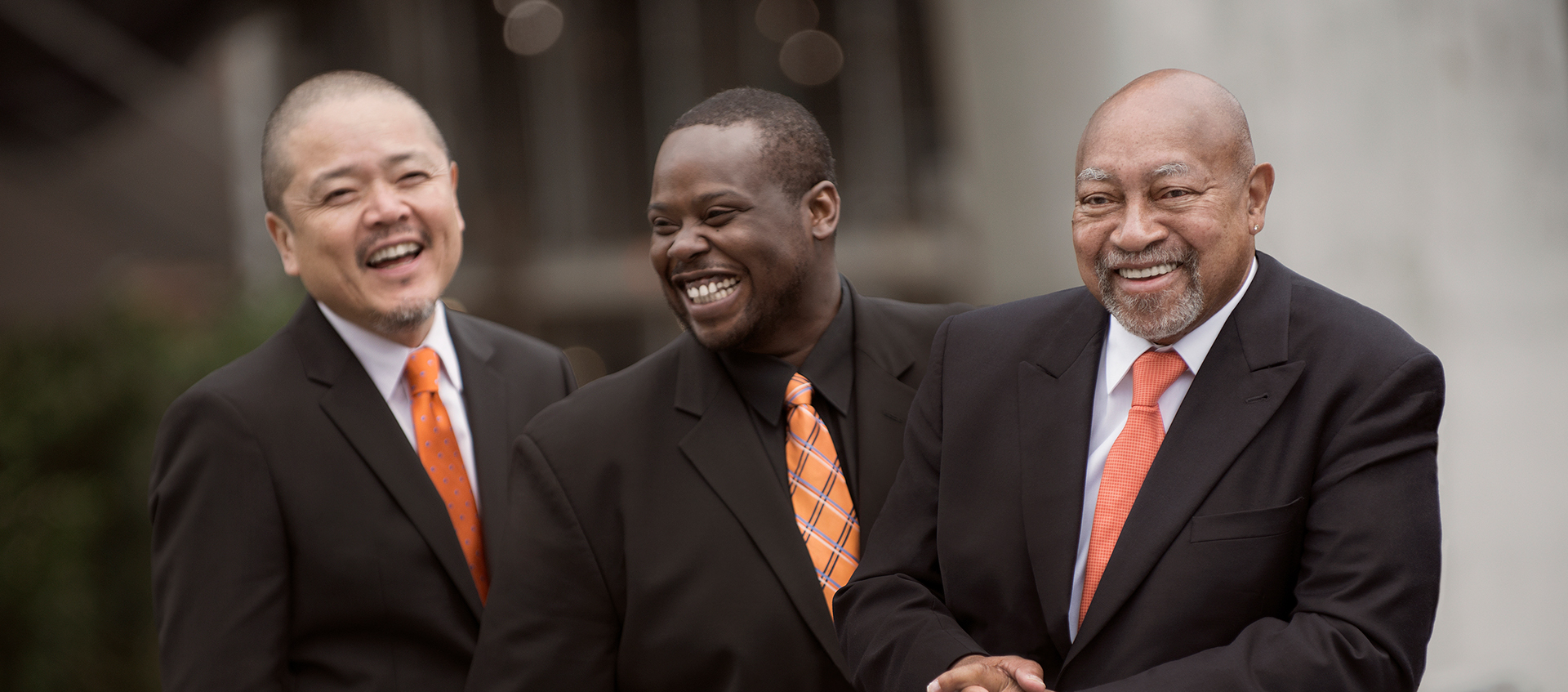 Three men in black suits and orange ties laugh together.
