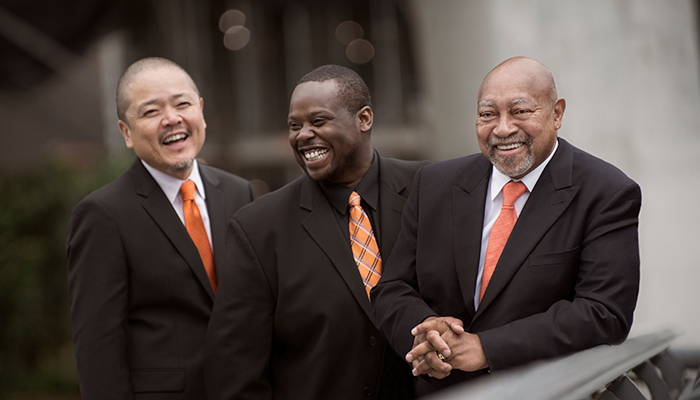 Three men in black suits and orange ties laugh together.