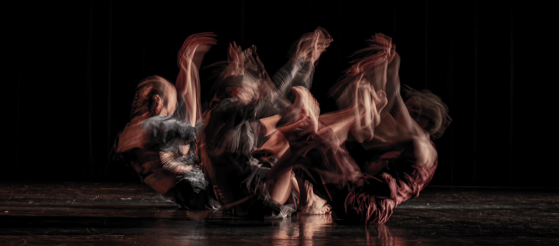 a blurred image of three dancers sitting on the floor with their hands raised.