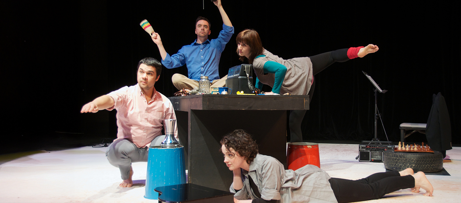 Two actors dance on top of a table while a third sits beside it and another lays beneath it.