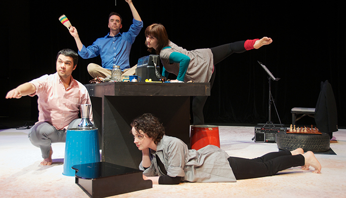 Two actors dance on top of a table while a third sits beside it and another lays beneath it.