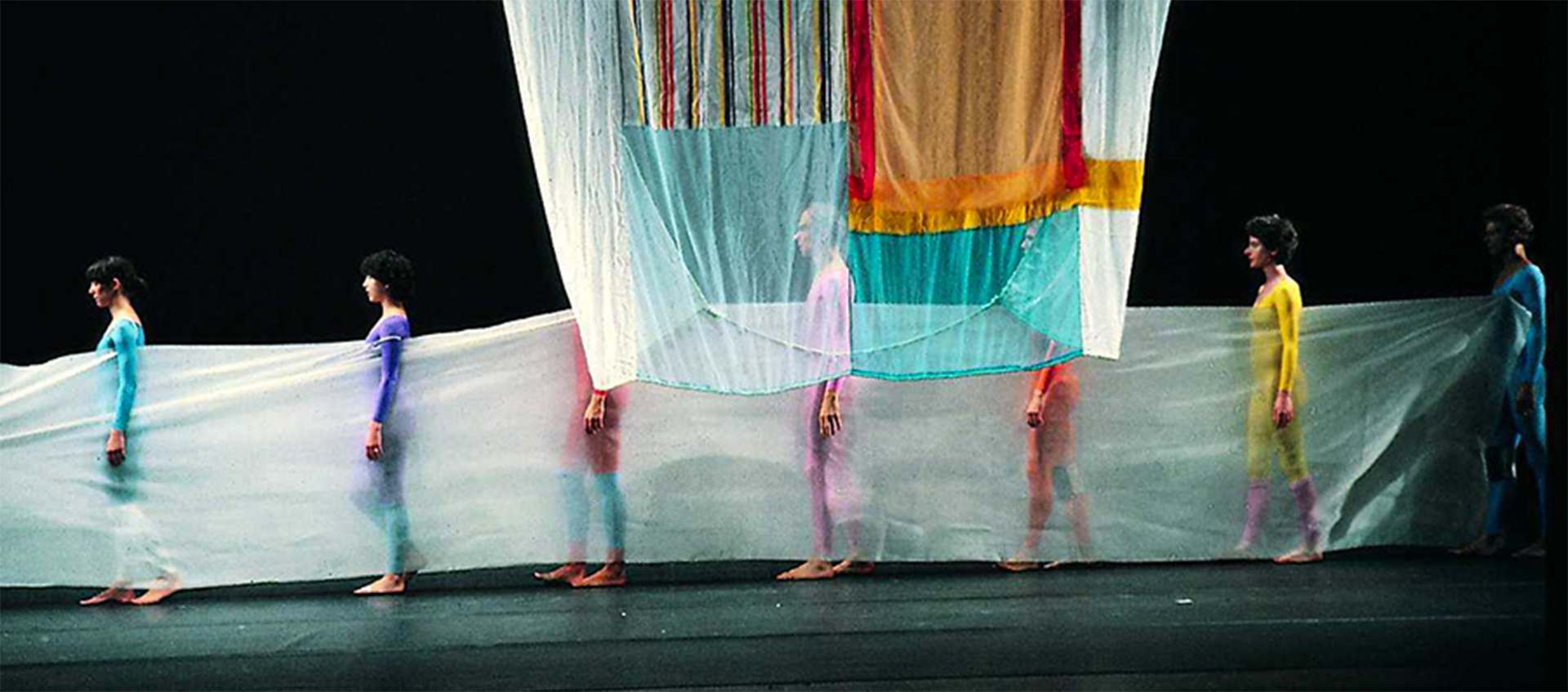 Dancers in colorful leotards stand behind a translucent cloth, a large cloth hangs from the ceiling in front of them.