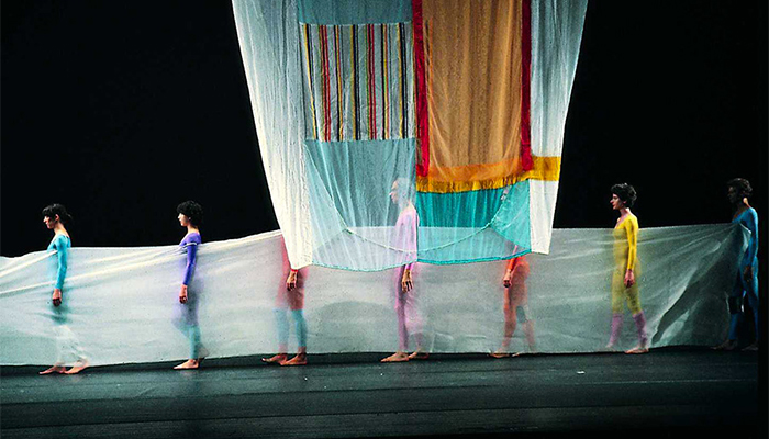 Dancers in colorful leotards stand behind a translucent cloth, a large cloth hangs from the ceiling in front of them.