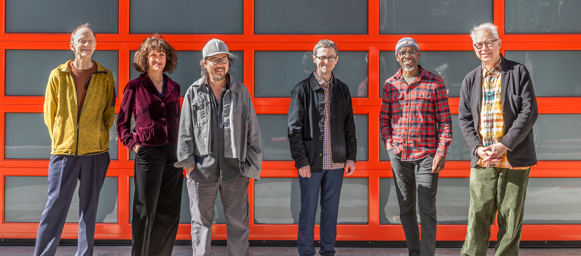 Six individuals stand together in front of a garage door with a grid of windows with orange trim.