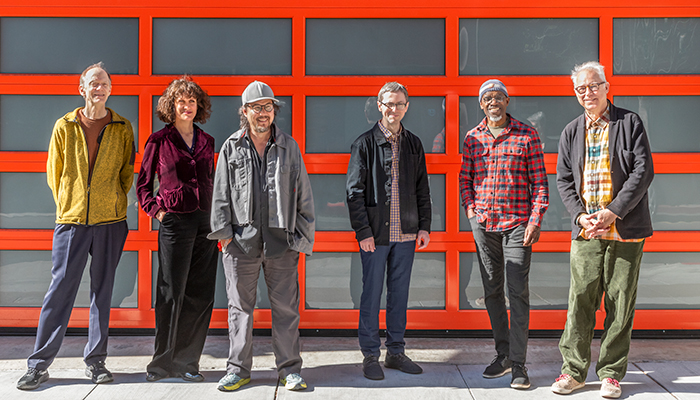 Six individuals stand together in front of a garage door with a grid of windows with orange trim.