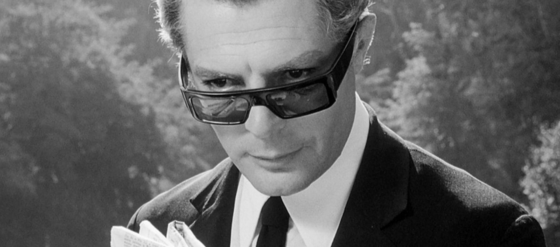 A black-and-white image of a man peeking over his sunglasses and looking slightly off-screen.