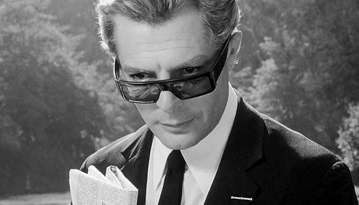 A black-and-white image of a man peeking over his sunglasses and looking slightly off-screen.