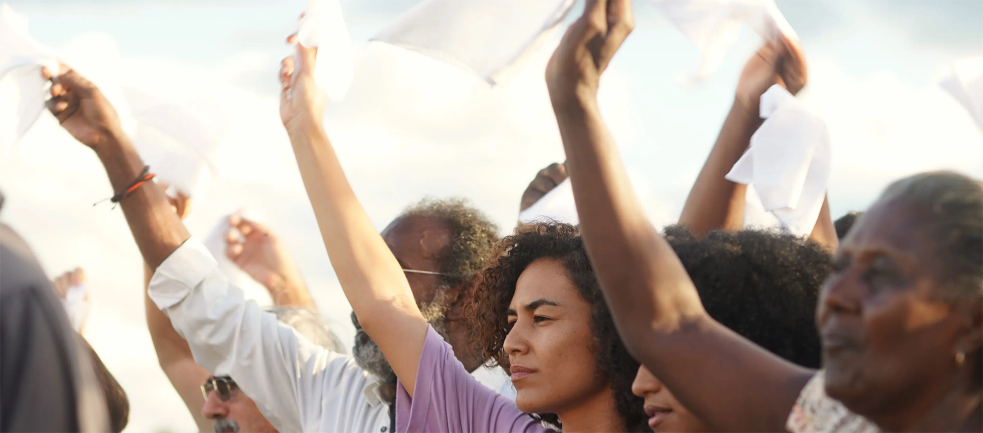 A group of people with a variety of hair colors, hair textures, and skin tones stand together and wave white handkerchiefs.