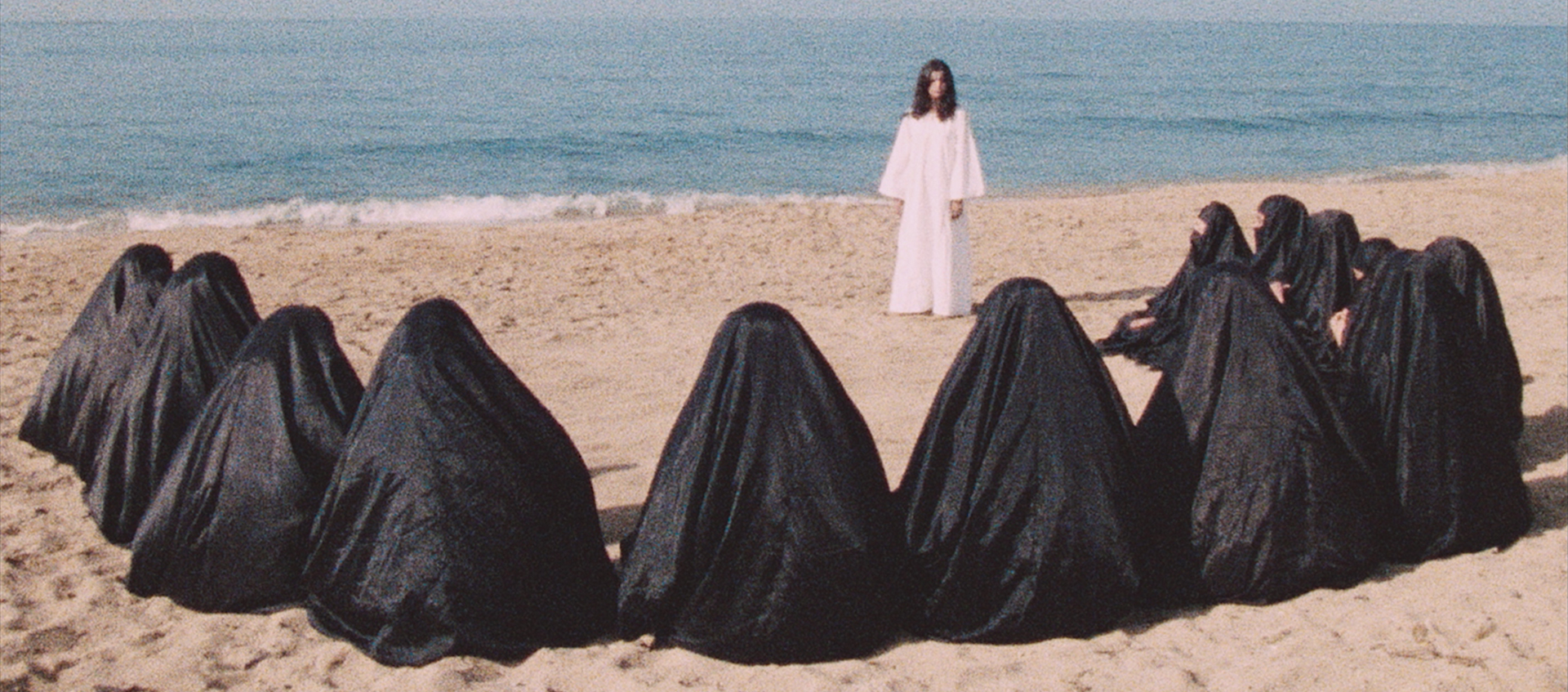 A woman wearing white stands in front of a group of people wearing black niqabs on the beach.