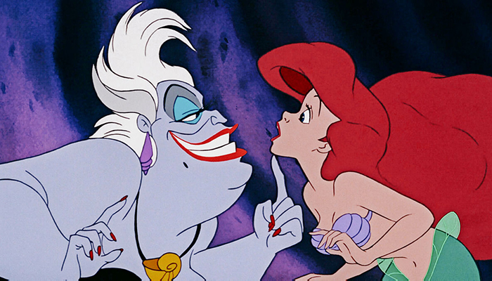 The purple-skinned sea witch Ursula tickles mermaid Ariel’s chin. Ariel has bright red hair.