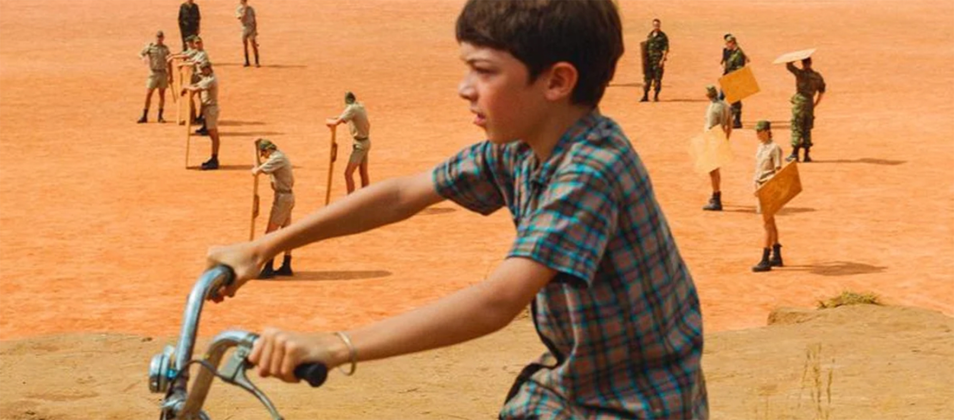 A young boy rides a bike. In the background, people in various military outfits stand around holding different objects.