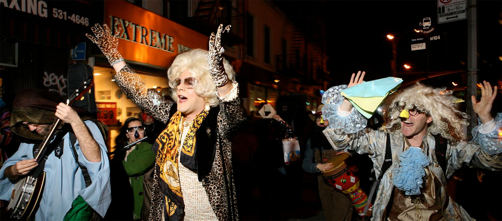 A drag queen in leopard print outfit raises her hands over her head. She is surrounded by a group of people in costumes.