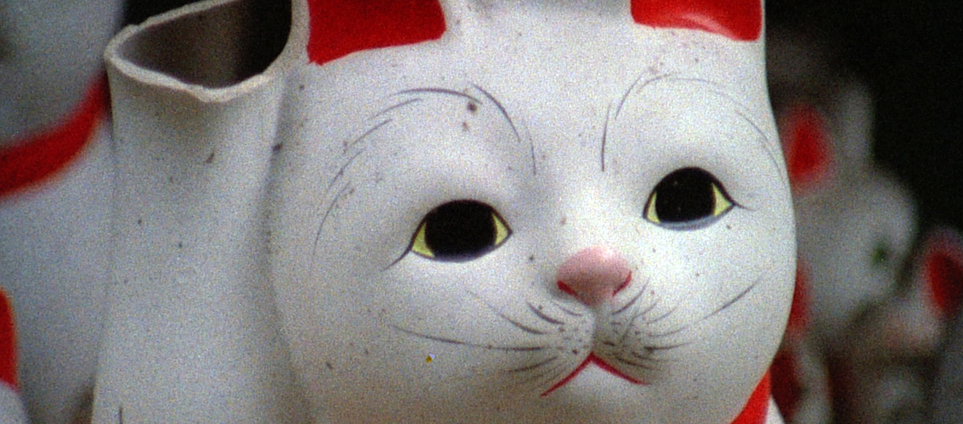 A closeup of a white cat sculpture with painted red ears, a red collar, and whiskers. The end of its raised paw is broken off.