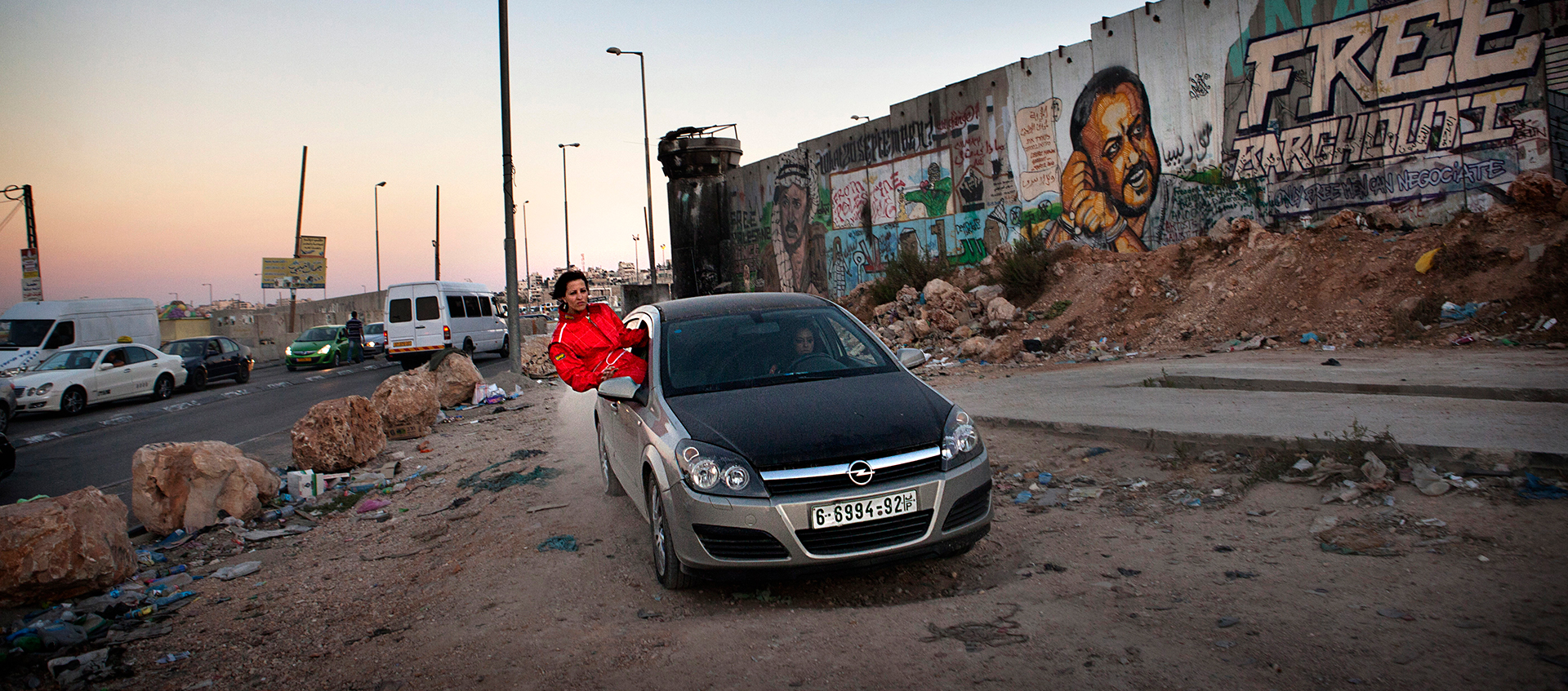 A person hangs out of the passenger seat of a car that is on a road next to a wall covered with graffiti.