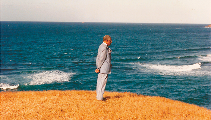A man in a gray suit stands on a grassy cliff overlooking the ocean, with waves and blue water visible in the background.
