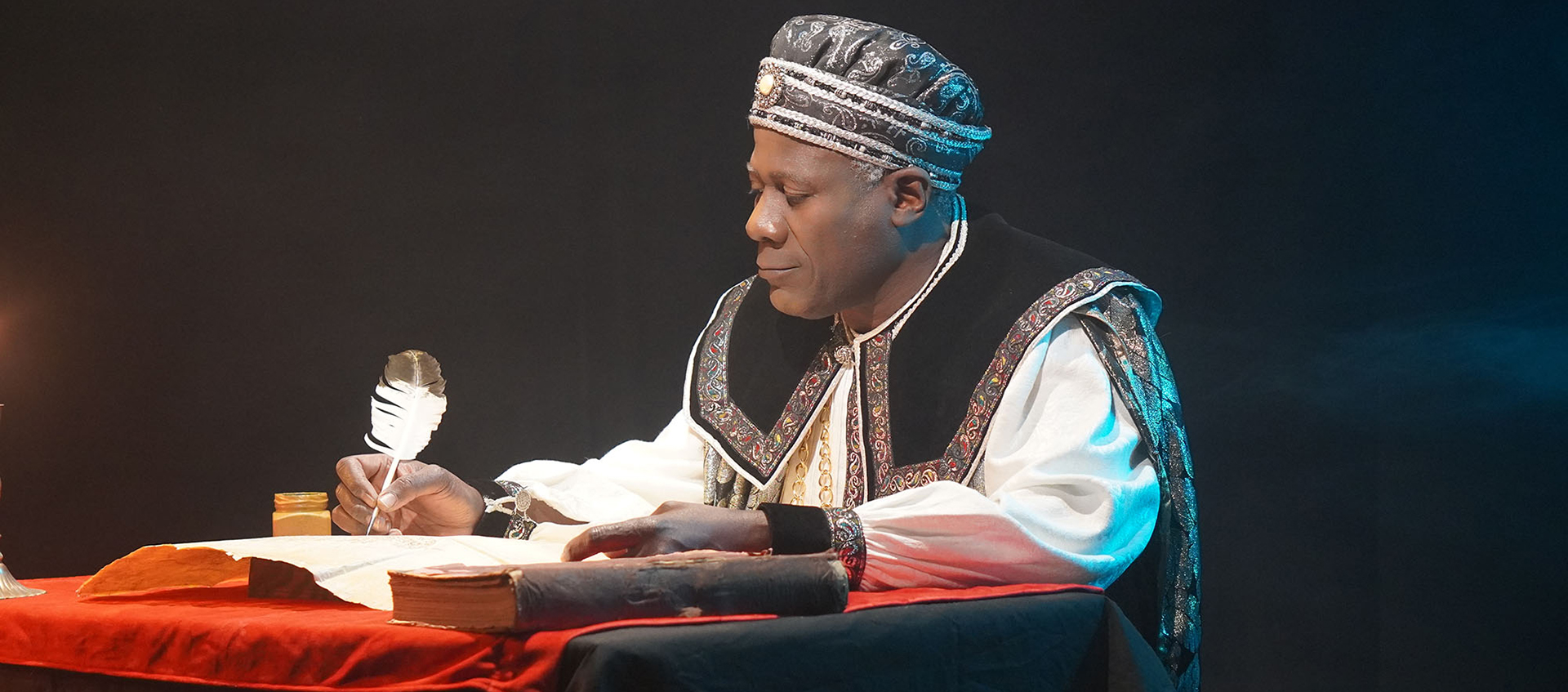 A Black man wears a regal-looking outfit and holds a quill in his hand as he writes on parchment.