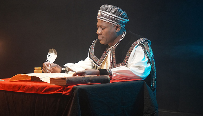 A Black man wears a regal-looking outfit and holds a quill in his hand as he writes on parchment.