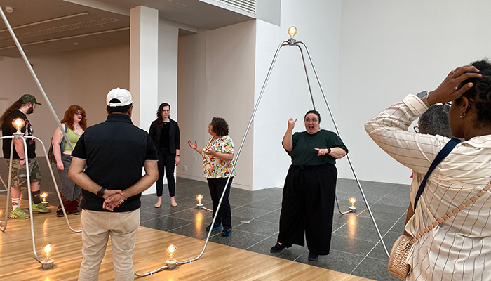 A group of people gather around an ASL interpreter in a gallery space featuring artwork from Nancy Holt: Power Systems.