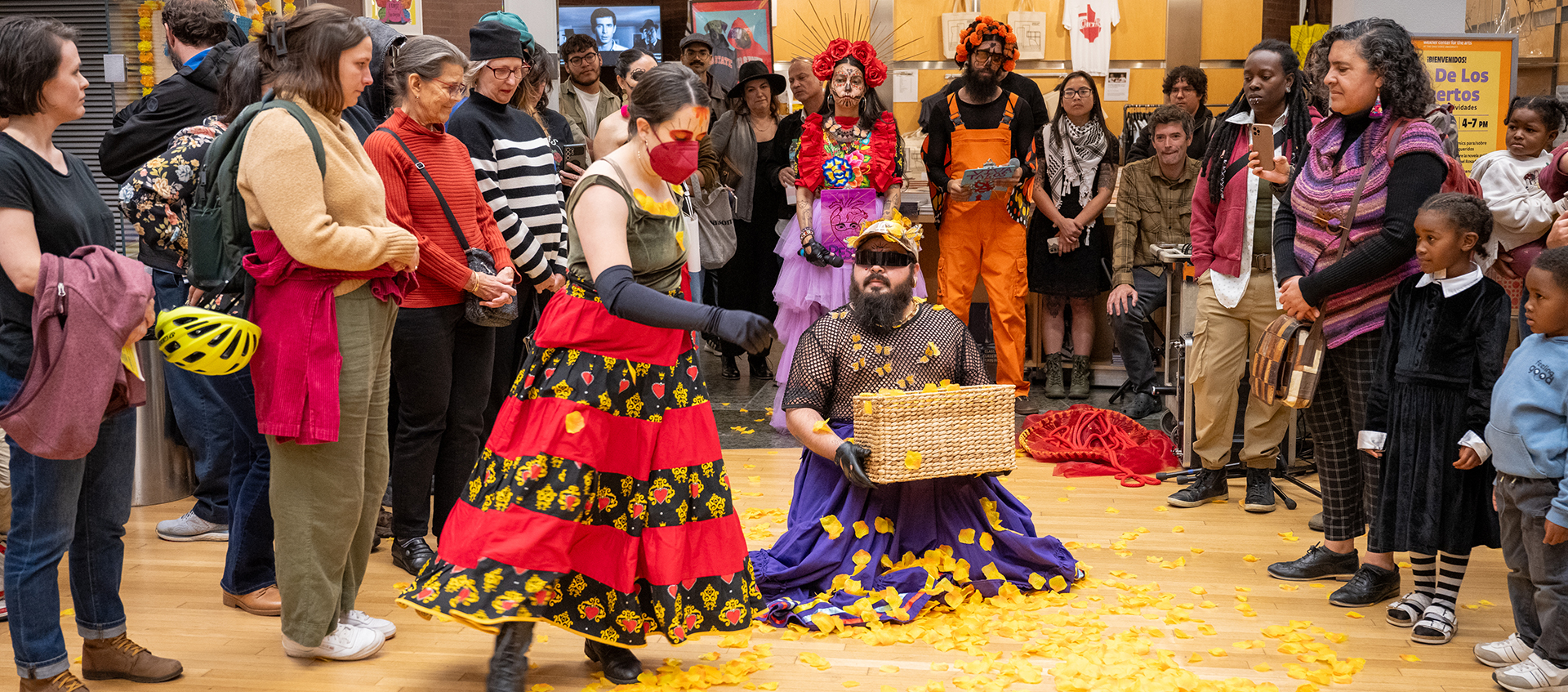 A crowd gathers in the Wexner Center around two performers wearing long, bright-colored skirts.