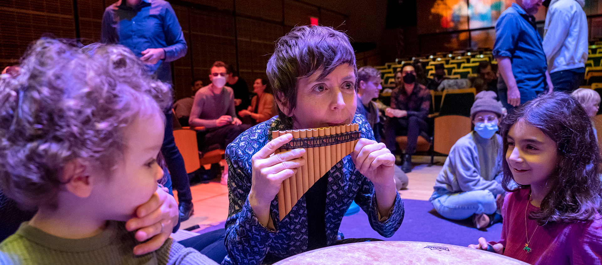 Claire Chase plays a pan flute on the floor of an auditorium. Two children sit next to her.