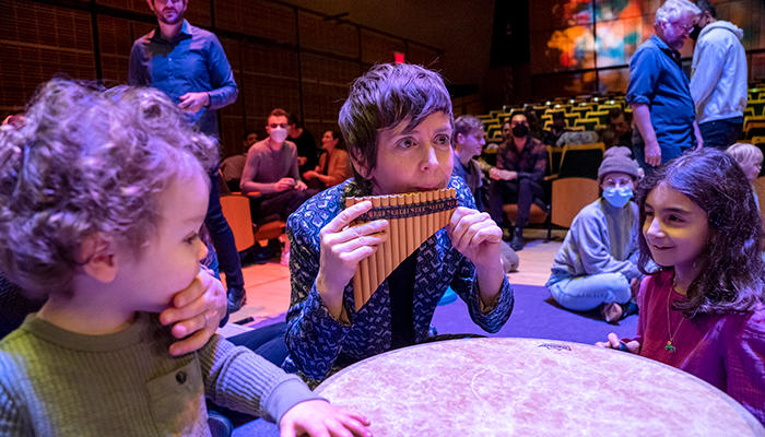 Claire Chase plays a pan flute on the floor of an auditorium. Two children sit next to her.