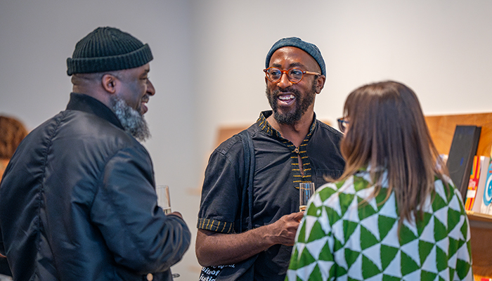 Three people stand in a group in an a gallery, smiling and talking while holding drinks.