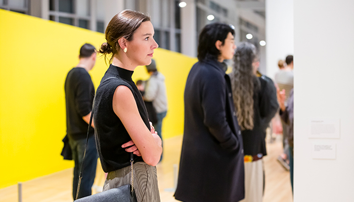 A woman stands with folded arms in a gallery, looking at artwork, with other visitors and a bright yellow wall in the background.