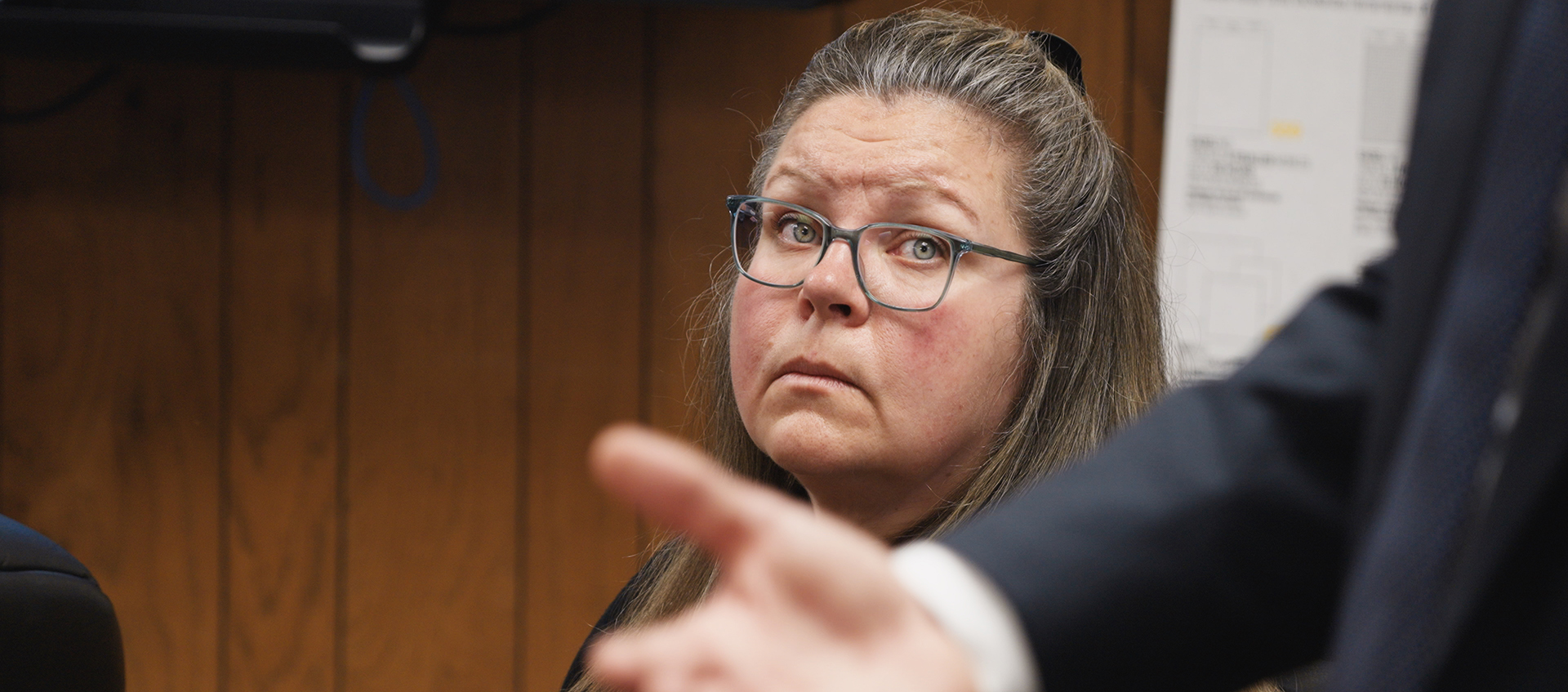 Closeup of a white woman with green eyes, graying brown hair, and glasses. She looks intently at a man beyond view.